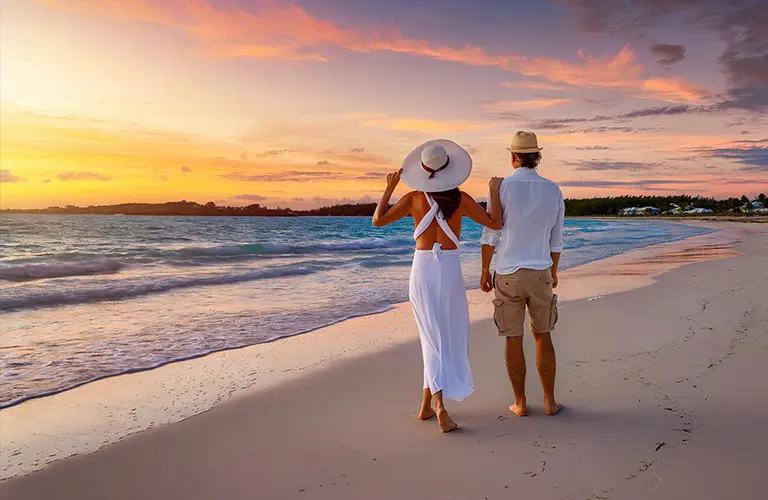 A couple wearing hats and white summer clothing standing on a tranquil sandy beach looking out at a vibrant sunset, a relaxing getaway organized by our Coastal Romance -  A 7 Day Boutique City & Sea Escape tour package
