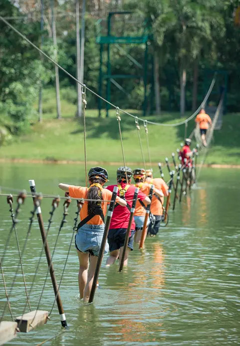 A group of people wearing safety helmets navigating a low rope bridge over a lake, highlighting the thrilling outdoor team building activities included in Corporate Showcase package