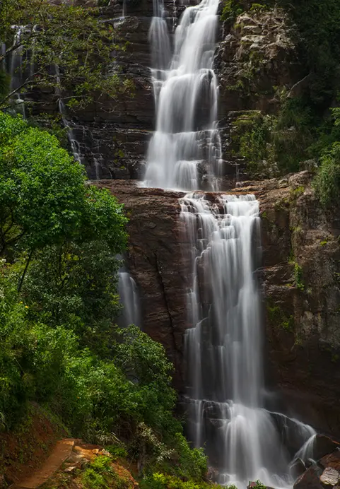 A stunning multi-tiered waterfall cascading down dark rocky cliffs surrounded by lush green foliage, a breathtaking nature stop frequently featured in our Magical Sri Lanka - The Ultimate 15 Day Family Adventure package