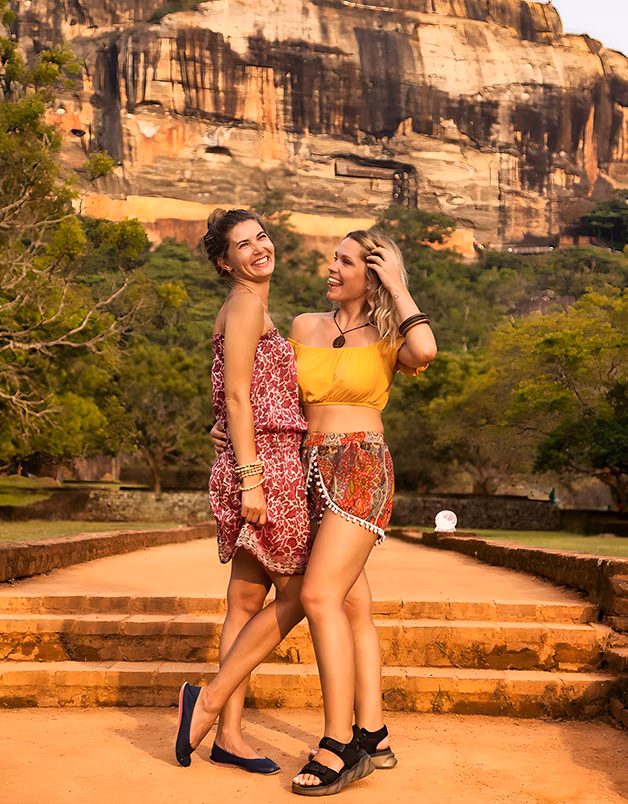 Two female friends laughing together on a pathway leading to the iconic Sigiriya Rock Fortress, enjoying a sunny day on their Sri Lanka cultural tour package