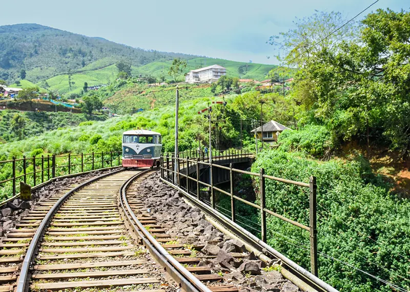 A vintage blue train travels along a curved track through green mountains