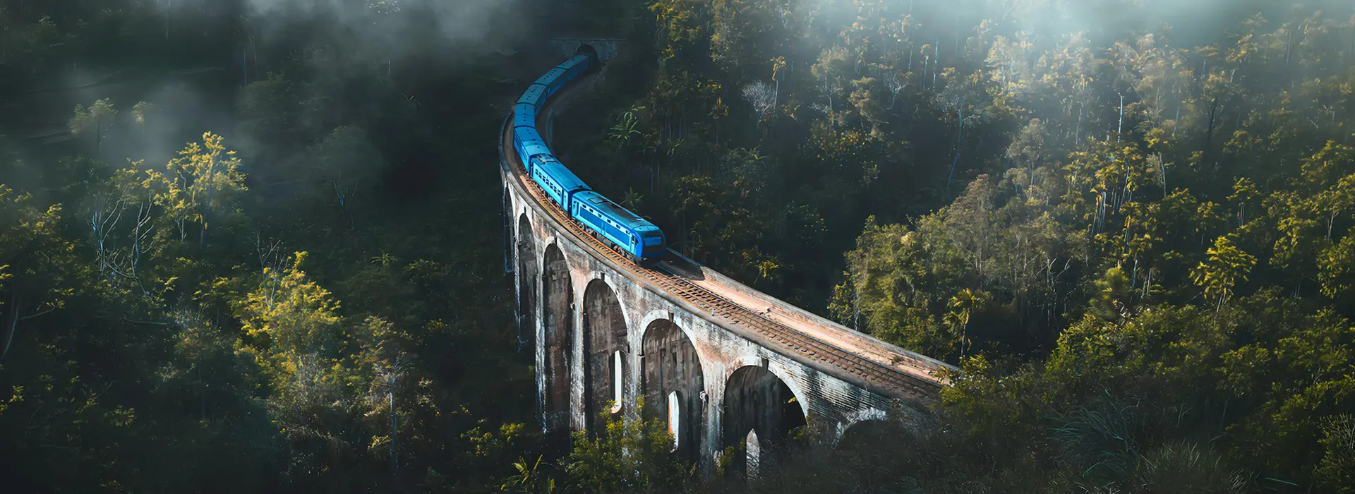 A scenic, panoramic view of the famous blue train crossing a historic arched viaduct surrounded by mist and tropical forest, highlighting the iconic experiences featured in our Sri Lanka tour packages