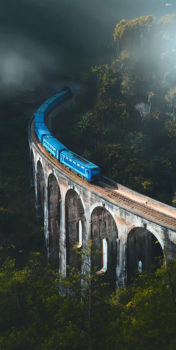 A scenic, panoramic view of the famous blue train crossing a historic arched viaduct surrounded by mist and tropical forest, highlighting the iconic experiences featured in our Sri Lanka tour packages