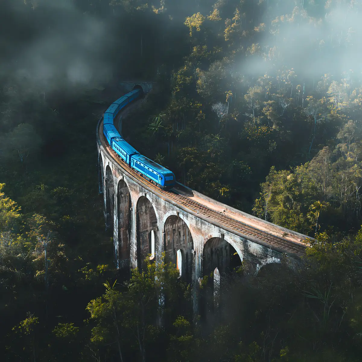 A scenic, panoramic view of the famous blue train crossing a historic arched viaduct surrounded by mist and tropical forest, highlighting the iconic experiences featured in our Sri Lanka tour packages