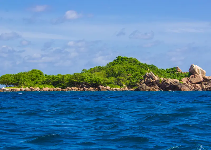 A small, lush green island with rocky shores surrounded by deep blue ocean water under a partly cloudy sky, a beautiful coastal destination to explore on our private Sri Lanka tours