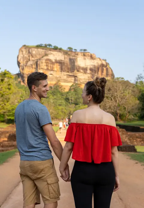 A smiling couple holding hands and walking along a dirt path towards the majestic Sigiriya Rock fortress during a Romantic Sri Lanka - A 12 Day Culture & Nature Odyssey tour