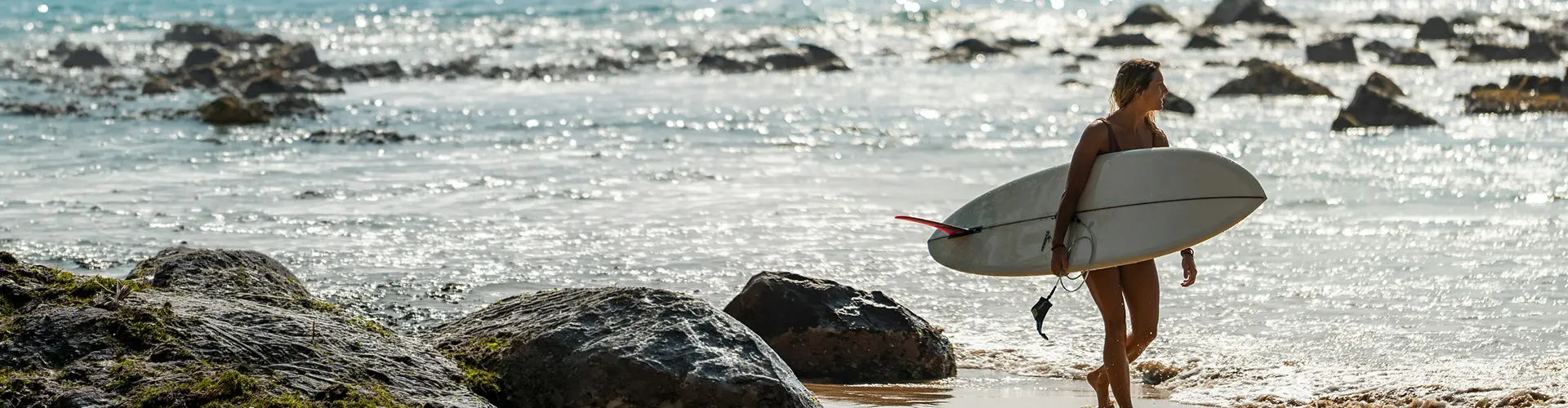 A female surfer carrying her white surfboard out of the sparkling ocean on a sunny beach, highlighting the exciting water sports and active coastal getaways included in our Sri Lanka Southern Sands Voyage package