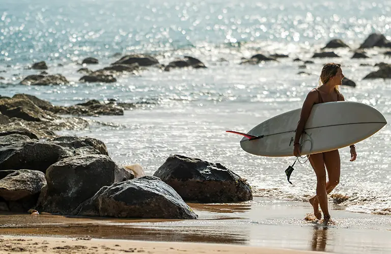 A female surfer carrying her white surfboard out of the sparkling ocean on a sunny beach, highlighting the exciting water sports and active coastal getaways included in our Sri Lanka Southern Sands Voyage package
