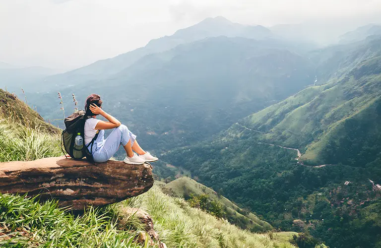 A female traveler with a backpack sits on a scenic rocky cliff overlooking a lush green valley, showcasing the incredible mountain hiking adventures included in our Sri Lanka adventure tour packages