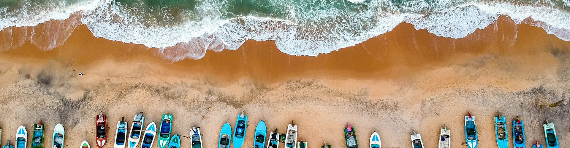 An aerial view of colorful fishing boats lined up on a sandy beach next to crashing ocean waves, highlighting the coastal destinations included in our Eastern Blues Tour.