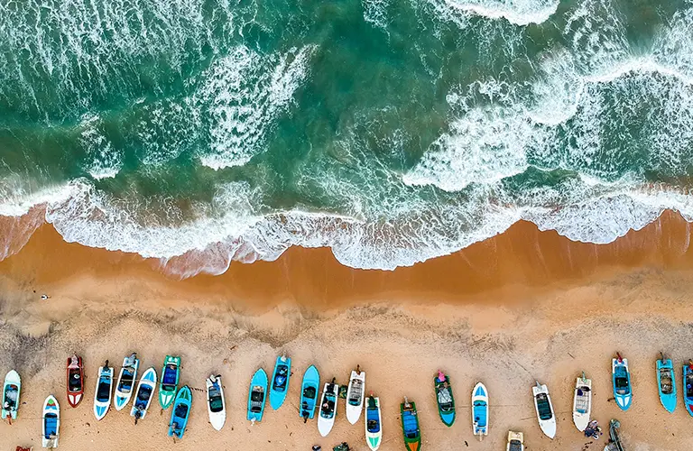 An aerial view of colorful fishing boats lined up on a sandy beach next to crashing ocean waves, highlighting the coastal destinations included in our Eastern Blues Tour.