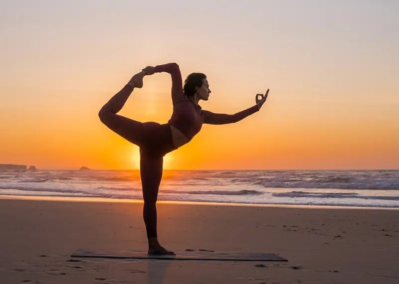 A woman practices a balancing yoga pose on a beach at sunset