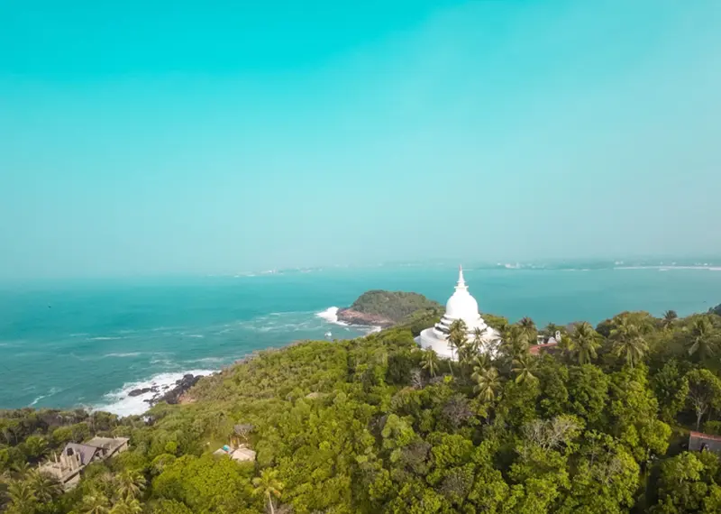 A white Buddhist stupa stands on a green hill overlooking the ocean