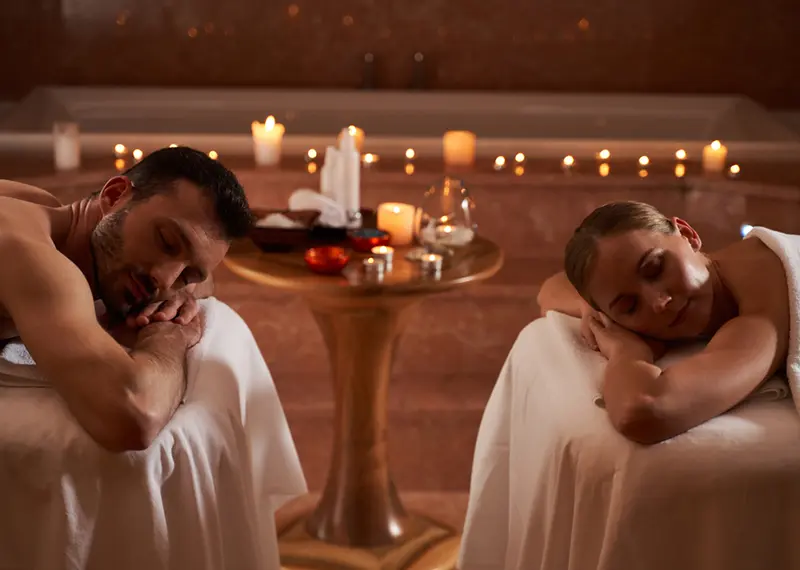 A couple relaxes on massage tables in a romantic candlelit spa setting