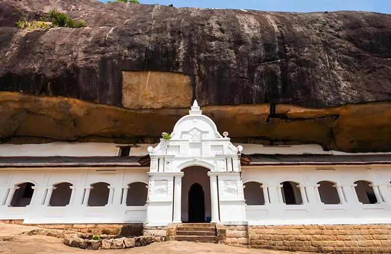The brilliant white arched entrance and veranda of the historic Dambulla Cave Temple built under a massive rock overhang, showcasing the ancient cultural sites included in our Sri Lanka The Kings & Kingdoms Express package