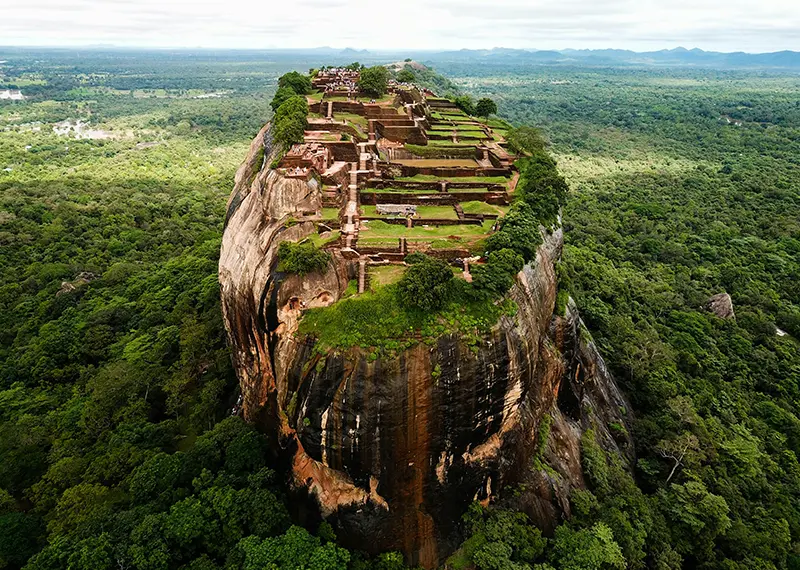 A stunning aerial view of the majestic Sigiriya Rock Fortress towering over a lush green jungle, showcasing ancient terraced ruins on its flat summit, a spectacular highlight included in our Sri Lanka tour packages from Australia