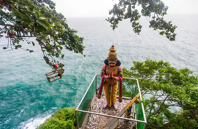 A colorful Hindu deity statue standing on a cliffside platform overlooking a vibrant turquoise ocean, framed by lush green trees, a breathtaking religious site featured on The Grand Ramayana & Heritage Expedition tour