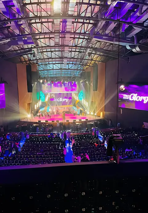 A wide view of a large, modern auditorium illuminated with purple stage lights and large screens during a corporate presentation, showcasing the night service at the Miracle Dome which can be experienced in The Miracle Dome Spiritual Encounter tour