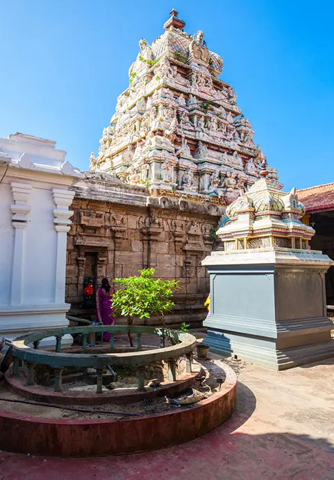 An intricately carved, multi-tiered Hindu temple tower under a bright blue sky, with a worshipper in a purple sari near a sacred courtyard enclosure, showcasing the rich cultural and religious sites included in our The Ramayana Trail package