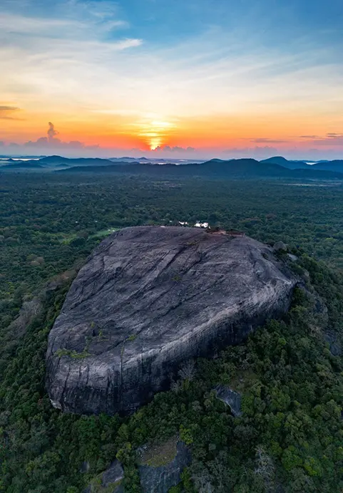 An aerial view of a massive rock formation surrounded by lush green jungle under a vibrant orange sunrise, highlighting the spectacular hiking destinations included in our Venture Further - The 11 Day Adventure & Wilderness Loop package