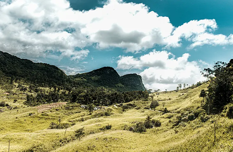 A picturesque view of a rural Sri Lankan valley surrounded by dramatic forested mountains and grassy slopes, representing the beautiful off-the-beaten-path destinations you can explore on Venture Further - The 11 Day Adventure & Wilderness Loop tours.