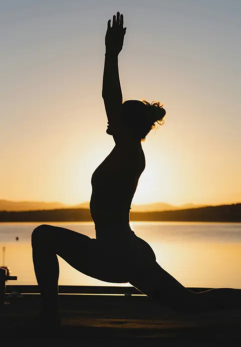 Silhouette of a woman practicing yoga by the water at sunset, a tranquil wellness experience featured in our tailored Wander & Awaken - The 11 Day Nervous System Reset package